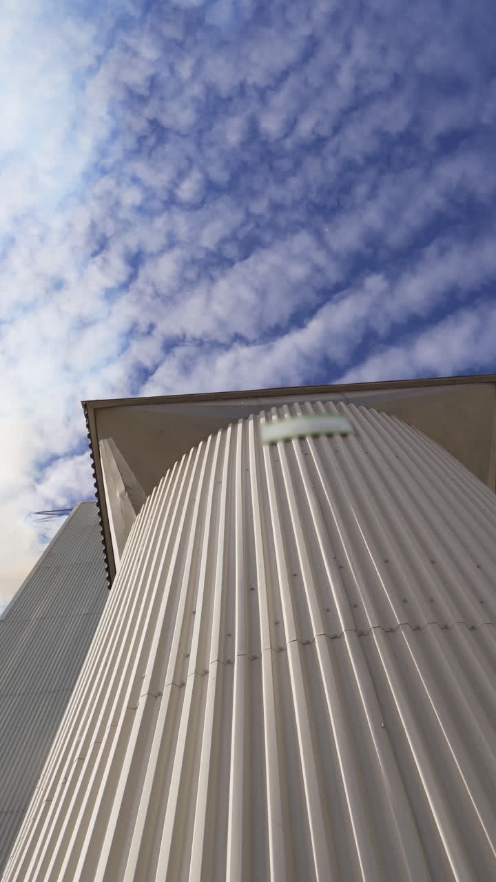 Metal grain elevator on sky background. Large granary for storing cereals. Thick smoke releasing into the air from industrial plant. Pollution. View from below. Vertical video