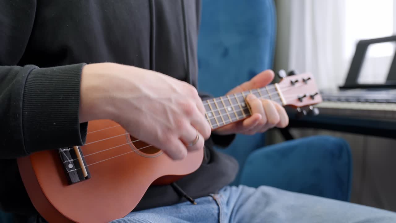 Musician is comfortably sitting on a blue armchair and playing ukulele, enjoying music and practicing chords in his apartment