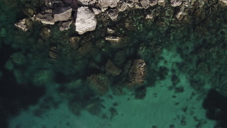 AERIAL---Turquoise-waters-give-way-to-rocky-cliffs-at-a-beach,-top-down-view