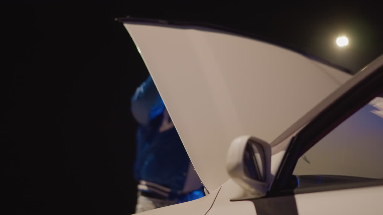 Driver examines motor under nighttime lamp, Male driver inspects vehicle engine on quiet deserted street, Individual assesses automobile engine beneath solitary streetlamp at nighttime