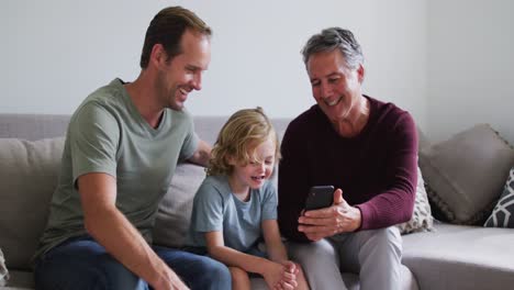 Abuelo-Caucásico,-Padre-E-Hijo-Sonriendo-Mientras-Usan-Un-Teléfono-Inteligente-Juntos-En-Casa