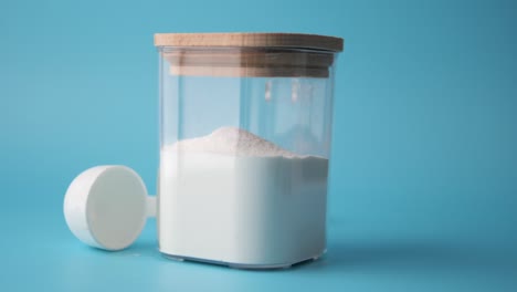 cerca de un frasco de vidrio lleno de polvo blanco, al lado de una cuchara de medición de plástico blanco sobre un fondo azul