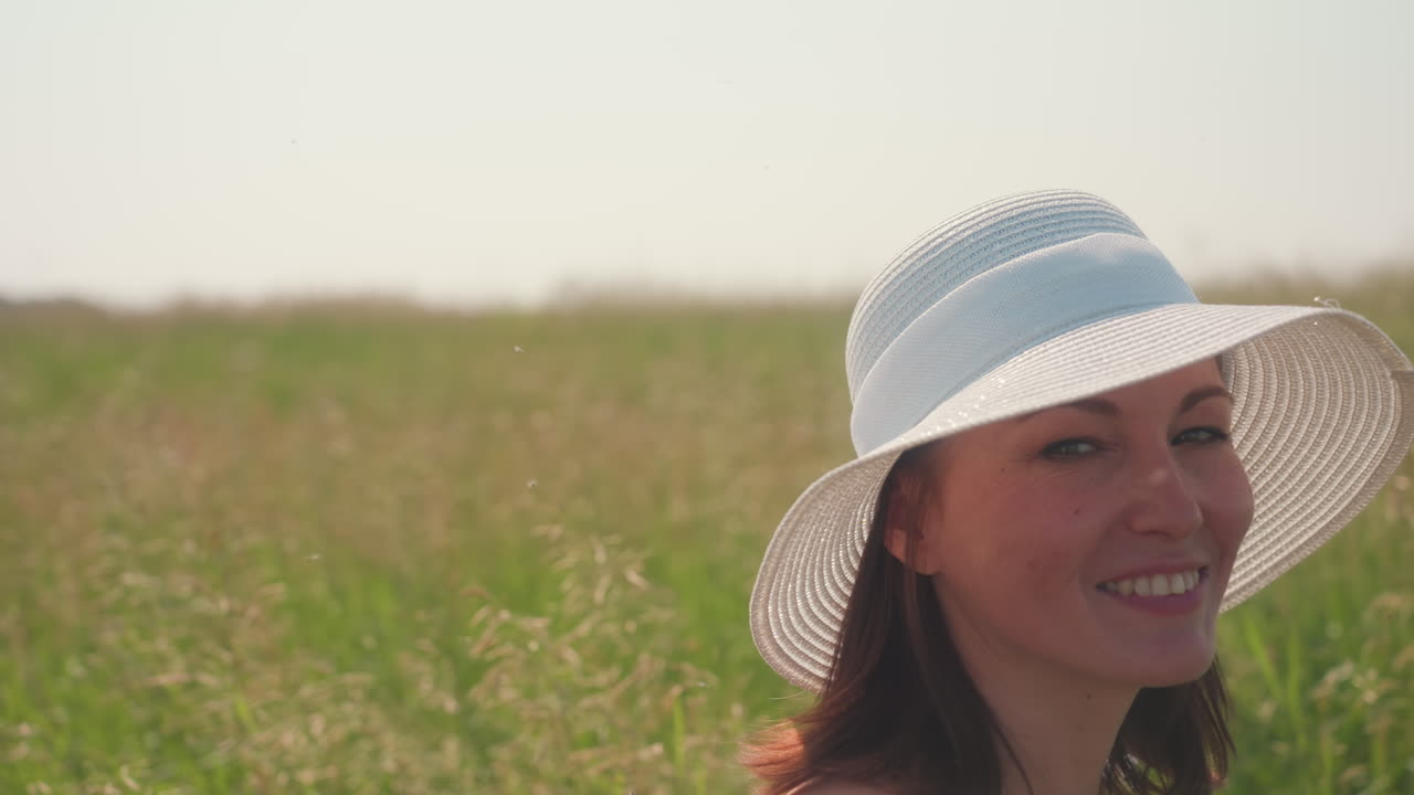 Close up of young woman smiling warmly at camera while wearing white sun hat in open barley field under clear summer sky, surrounded by flying insects and soft natural light