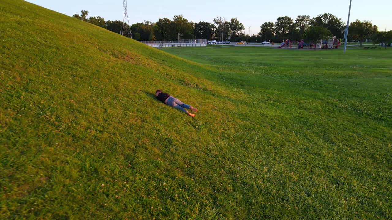 Happy boy rolls down green hill, aerial follow view