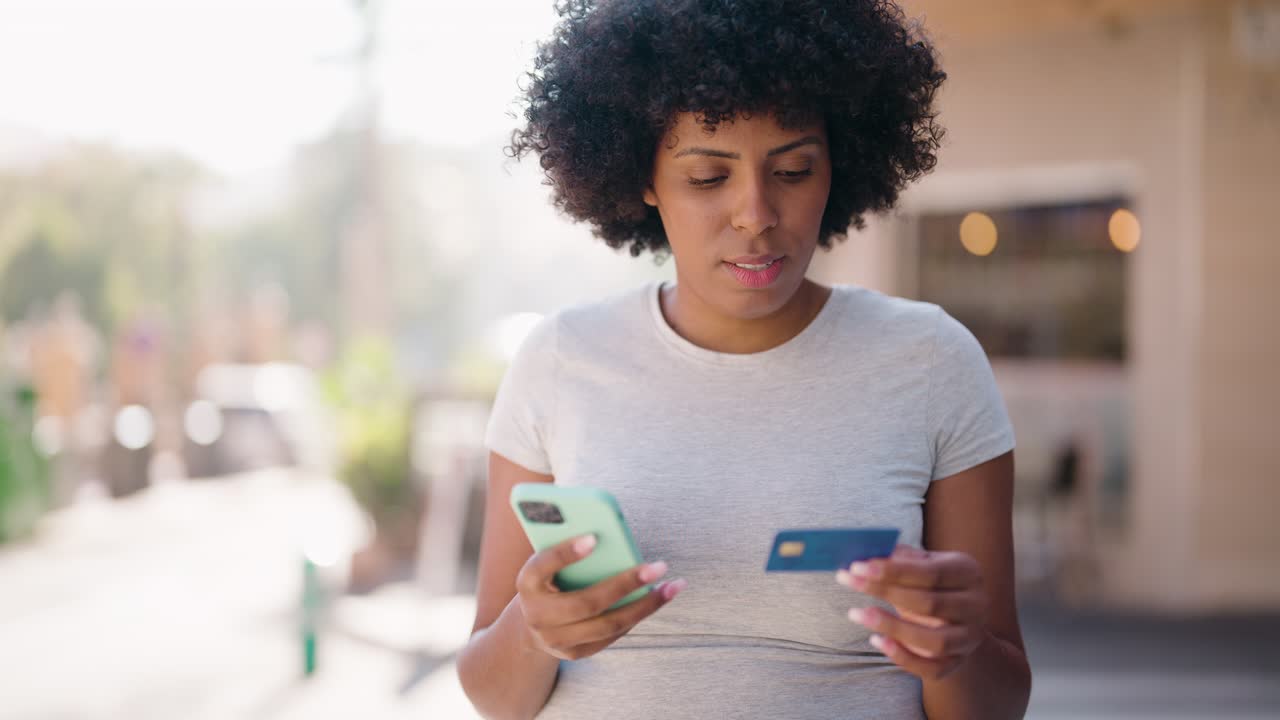 joven afroamericana usando teléfono inteligente y tarjeta de crédito en la calle