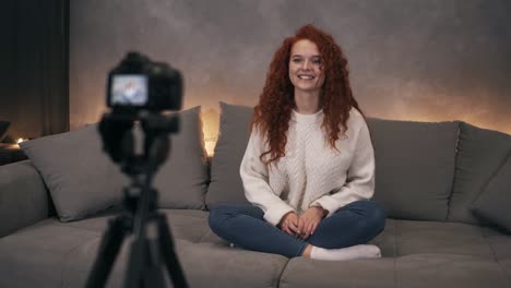 attractive curly red headed young girl vlogger is talking in front of camera recording video for online blog in internet speaking, smiling and gesturing. woman is wearing jeans and white sweater. accelerated video