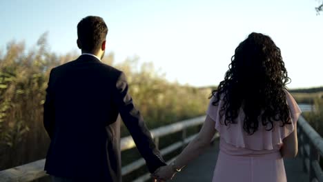 Slow-Motion-Back-view-of-engaged-couple-walking-hand-in-hand-on-wooden-walkway-in-park