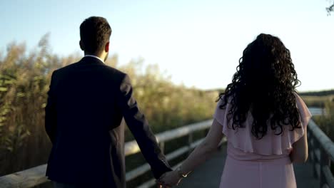 slow motion-back view of engaged couple walking hand in hand on wooden walkway in park