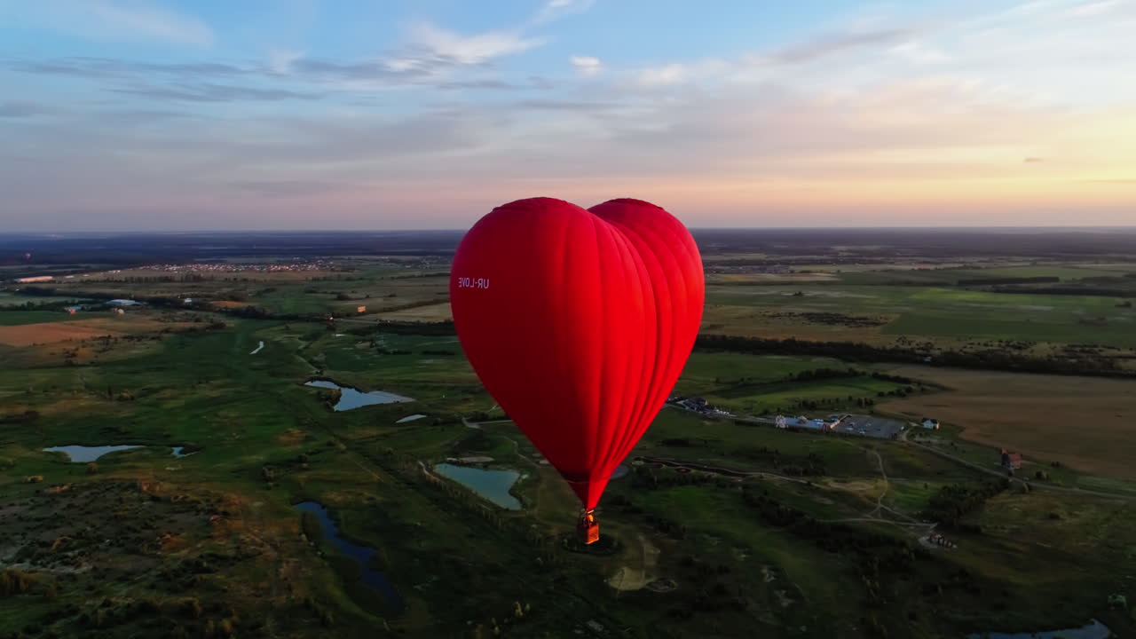 Aerostat in the hear form in the air. Red balloon fly over green meadows with lakes at sunset. Aerial view. Camera moving around.