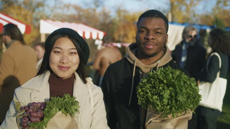 happy couple with goods at marketplace looking at camera diverse couple standing with paper bags of vegetables or fruits spouses looking at camera smiling people buy healthy and fresh food on background shopping at local farmers market portrait view