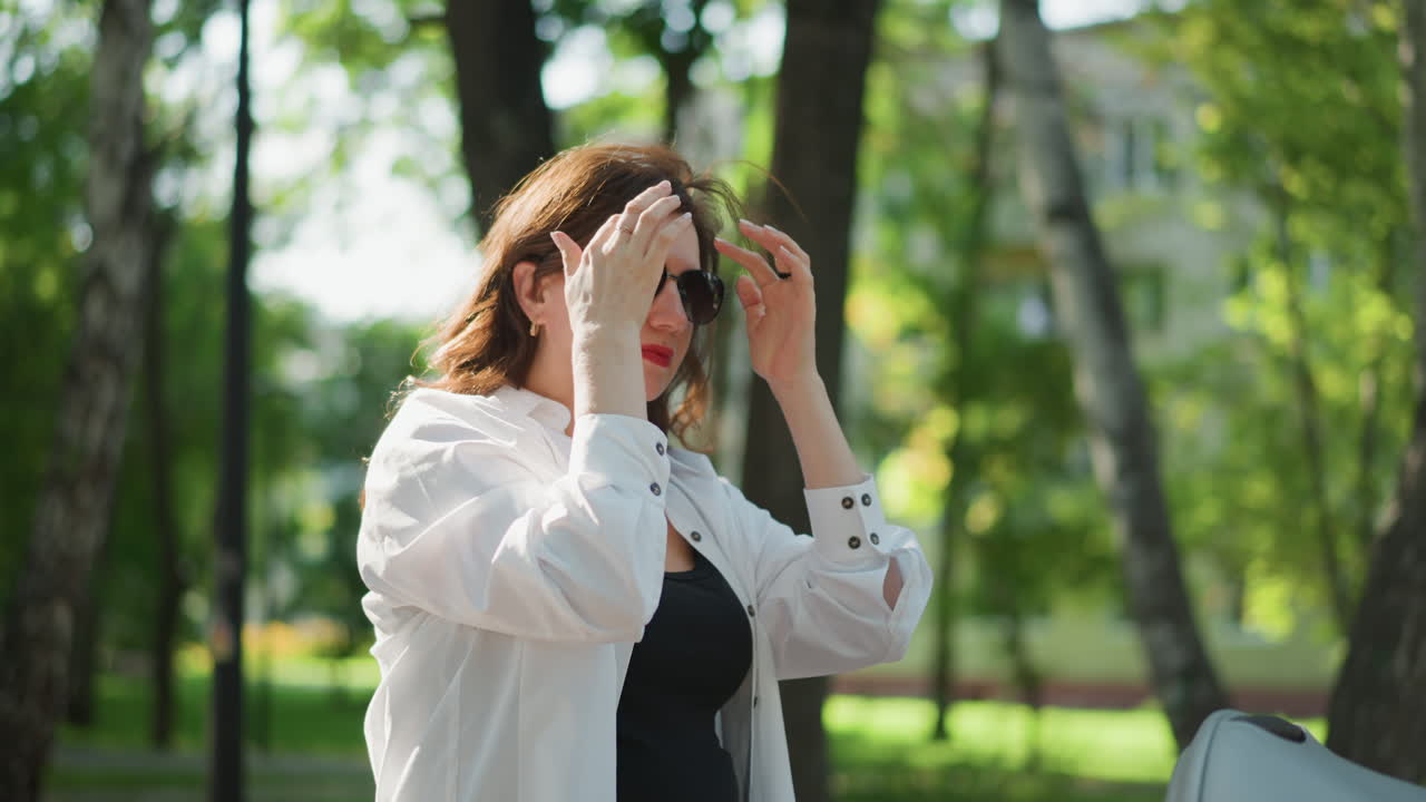 Donna bianca in un parco soleggiato che aggiusta gli occhiali da sole e riflette su un principio di vita personale, camicia bianca casual, collana, controluce caldo attraverso gli alberi, atmosfera serena del ritratto, rilassata e contemplativa.