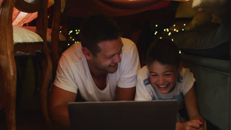 Padre-E-Hijo-Caucásicos-Sonriendo-Mientras-Usan-Una-Computadora-Portátil-Bajo-Una-Manta-Fuerte-Durante-La-Navidad-En-Casa