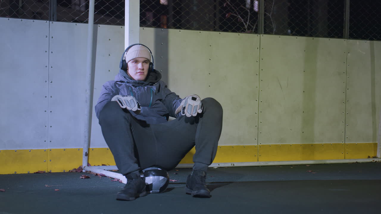 골 포스트에 기대어 땅에 앉아 있는 남자, 장갑, 후디, 헤드폰을 착용하고, 발 에 축구 공을 가지고, 체인 링크 울타리와 배경에 도시 조명으로 둘러싸여 있습니다.