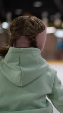 primer plano de una mujer patinando en una pista de hielo, con una sudadera verde menta y una cinta rosa con su cabello en trenzas