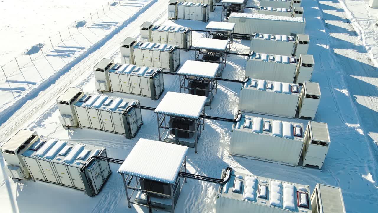 Low-altitude winter aerial rises above containerized battery units, revealing high-voltage infrastructure, snowy geometry, and the scale of Estonia’s flagship energy storage site