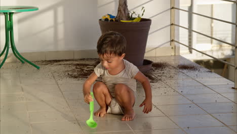 naughty little toddler playing with a green plastic spoon after soiling all the floor with dirt from a plant pot