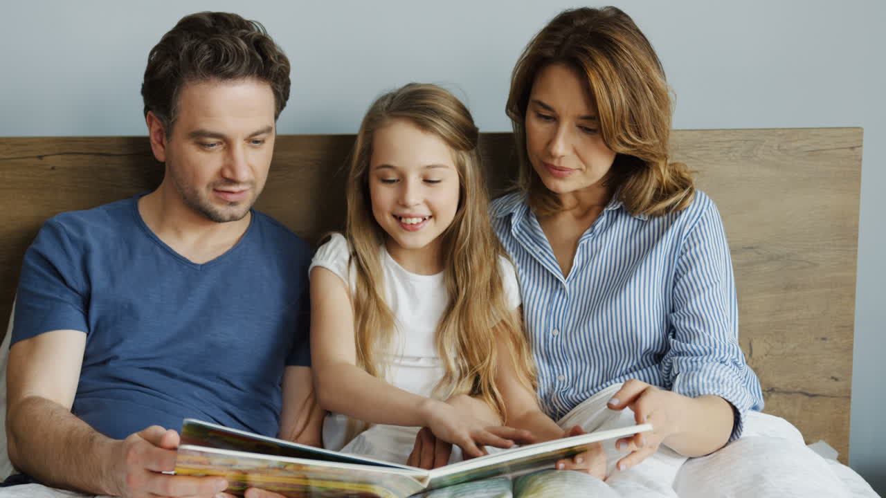 Young mother and father sitting on the bed in the morning with their little daughter and reading an interesting book