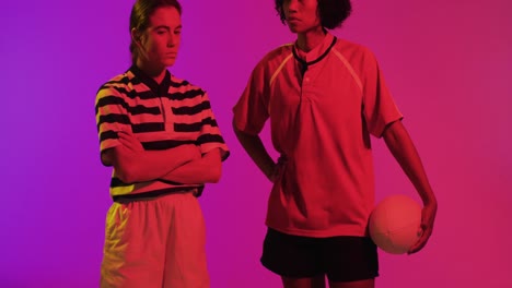 portrait of diverse female rugby players with rugby ball over neon pink lighting