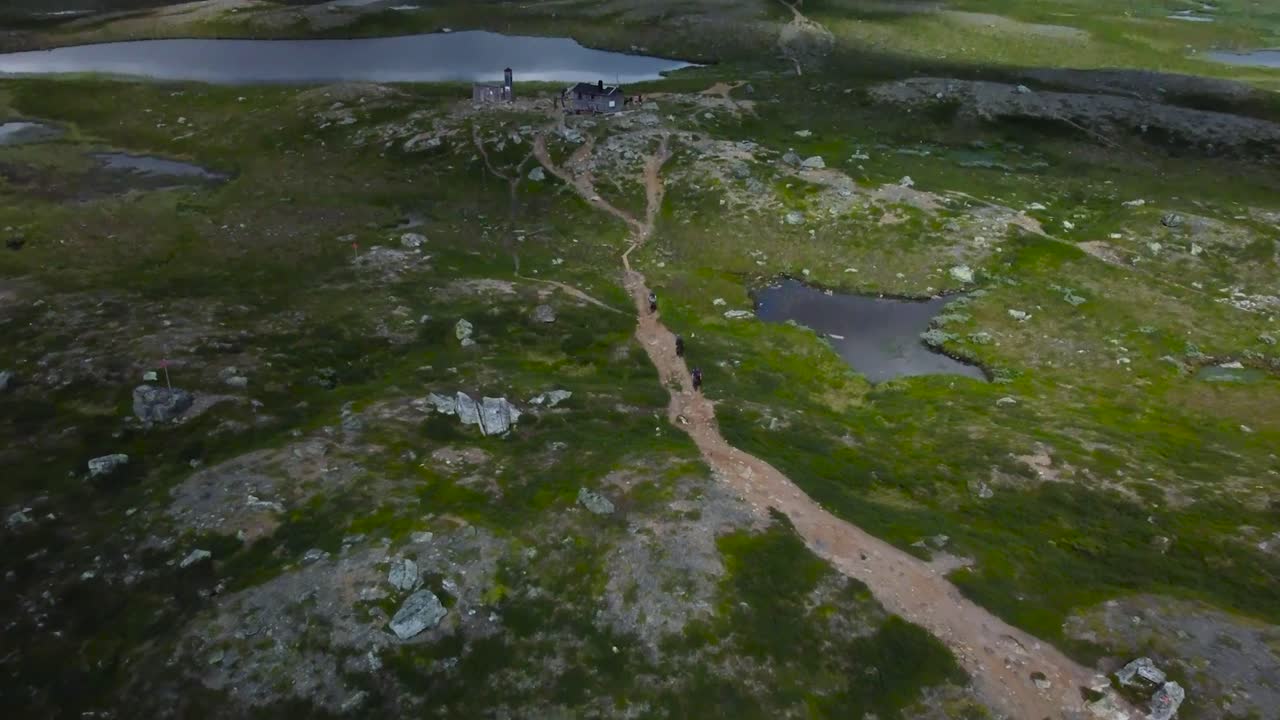 Aerial drone footage flying backwards and revealing people walking or hiking on muddy brown colored road in Sweden nature rocky terrain landscape with small water puddles that are reflecting at day.