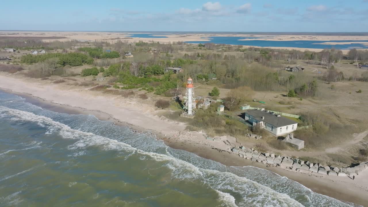 vista aérea del faro de color blanco, costa del mar báltico, letonia, playa de arena blanca, grandes olas chocando, día soleado con nubes, amplia toma de dron moviéndose hacia adelante, la cámara se inclina hacia abajo