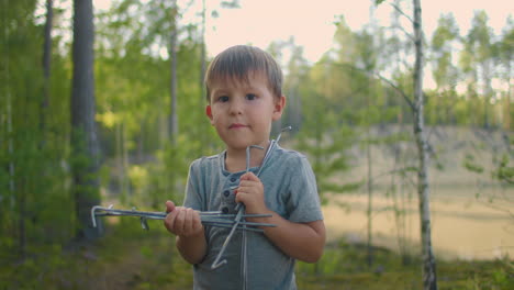 Un-Niño-De-3-Años-Mira-A-La-Cámara-Mientras-Está-Parado-En-El-Bosque-Y-Sostiene-Partes-De-Una-Tienda-De-Campaña-Para-Instalarla-Mientras-Acampa-En-Cámara-Lenta