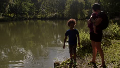 mother with a newborn in a baby carrier with child plays with water in city pond