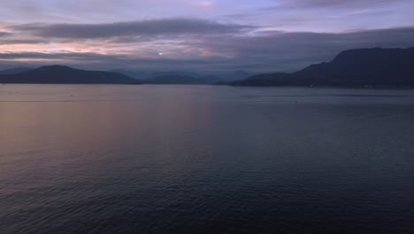 Aerial-Sunset-Wide-Shot-Flying-Over-Pacific-Ocean-Towards-Distant-Coastal-Mountain-Peaks-Of-Sunshine-Coast-Panning-Right-To-Reveal-North-Shore-And-Cargo-Ships-In-Vancouver-British-Columbia-Canada