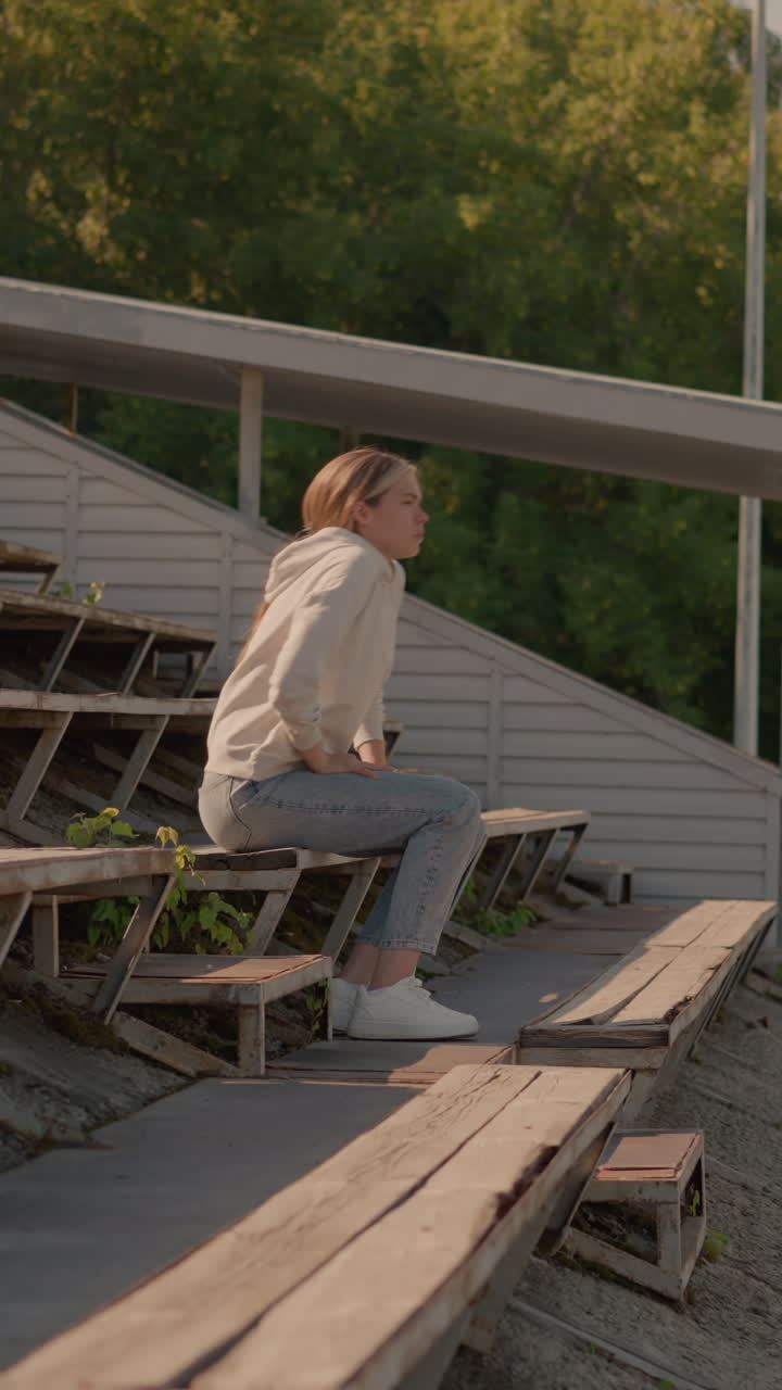 캐주얼 청바지와 후디를 입은 여자가 농촌 스타디움 좌석을 걸어가며 편안하고 반사적인 자세로 머리를 손에 고, 배경에는  빈 관중석, 전기 기둥, 활기찬 녹색 나무가 있습니다.