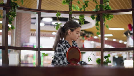 joven coloca un espejo de madera en la mesa y se mira a sí misma mientras ajusta el cabello con la mano, sentada en un espacio interior acogedor con decoración natural, parece enfocada y contemplativa
