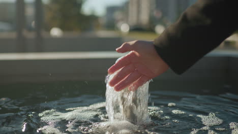cerca de la mano alcanzando el agua de manantial que fluye de una fuente con reflejos en la superficie del agua brillante, figura borrosa y fondo