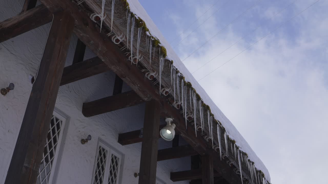 A mesmerizing sight unfolds as numerous glistening ice stalactites delicately hang from the roof's edge, gradually dissolving under the radiant rays of a sunny winter day