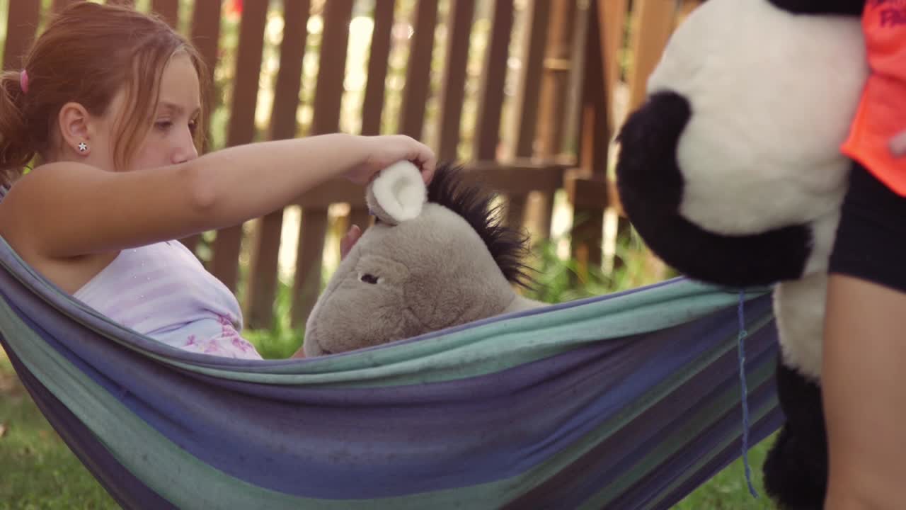 여름 캠프에서 행복한 어린 소녀들은 다채로운 햄에 누워서 밖에서 플러시와 함께 놀고, 초록 잔디에서 개 인형과 함께 놀고 있습니다.