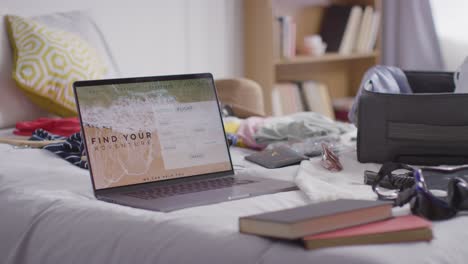 maleta abierta en la cama en casa siendo empacada para las vacaciones de verano con una computadora portátil que muestra los detalles de reserva de vuelos y hotel 2
