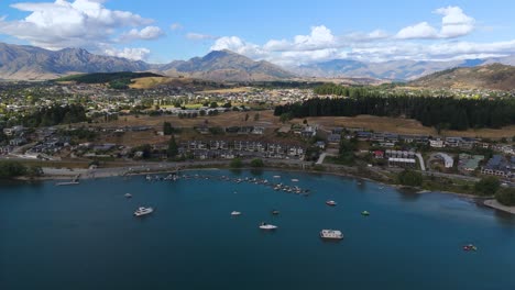 Frente-Al-Lago-Wanaka,-Barcos-Amarrados-En-Aguas-Azules-Con-Un-Telón-De-Fondo-De-Montañas-Y-Ciudad,-Vista-Aérea