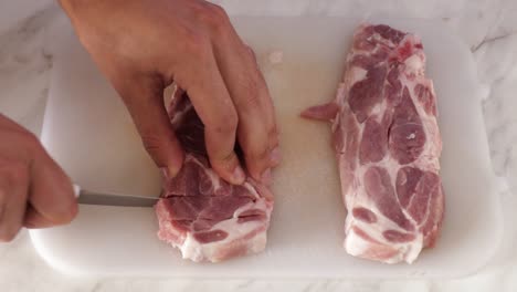 Closeup-of-a-mans-hand-cutting-pork-chops-on-a-white-board-with-a-steel-knife