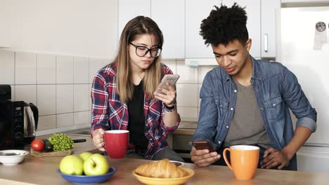 Cute-couple-having-breakfast-together-at-home-in-their-kitchen,-attractive-caucasian-girl-and-her-mulatto-boyfriend-using-their-smartphones,-not-talking-to-each-other-at-first.