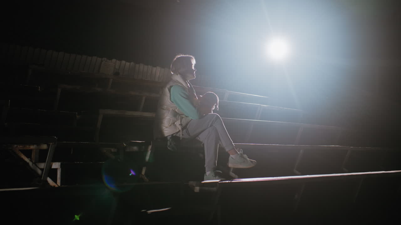Bright light shining over football stadium seats as elegant lady sits with her puppy in calm, serene moment during night, showcasing companionship, and loyalty bond under empty illuminated stands