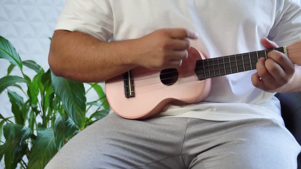 hombre de mediana edad 30 y con cuentas está jugando y silbando con su ukulele rosado al lado de una gran ventana y cerca de una planta verde