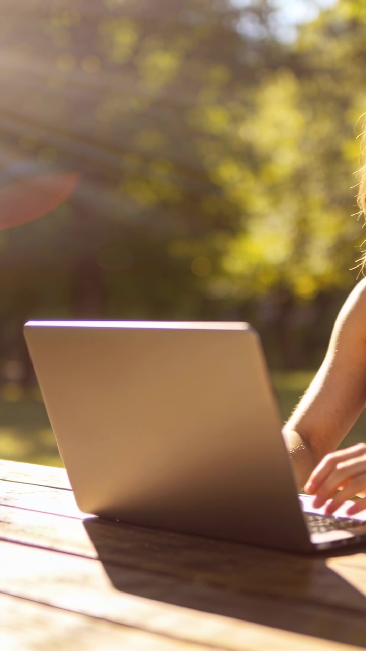 Video verticale: Donna con top senza maniche che si protende, digitando sul laptop a un tavolo da picnic, lavorando all'aperto.