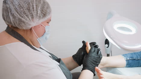technician wearing protective mask, hair cover, and black gloves seen massaging underside of client foot under bright lamp in clean, well lit salon setting. client is lying on chair in relaxed posture