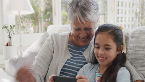 Chica-Feliz-Usando-Un-Teléfono-Inteligente-Con-Una-Abuela-Teniendo-Un-Chat-De-Video-Saludando-A-Una-Familia-Compartiendo-El-Fin-De-Semana-De-Vacaciones-Con-Una-Abuela-Charlando-Por-Teléfono-Móvil-Relajándose-En-Casa-Con-Su-Nieta-4k