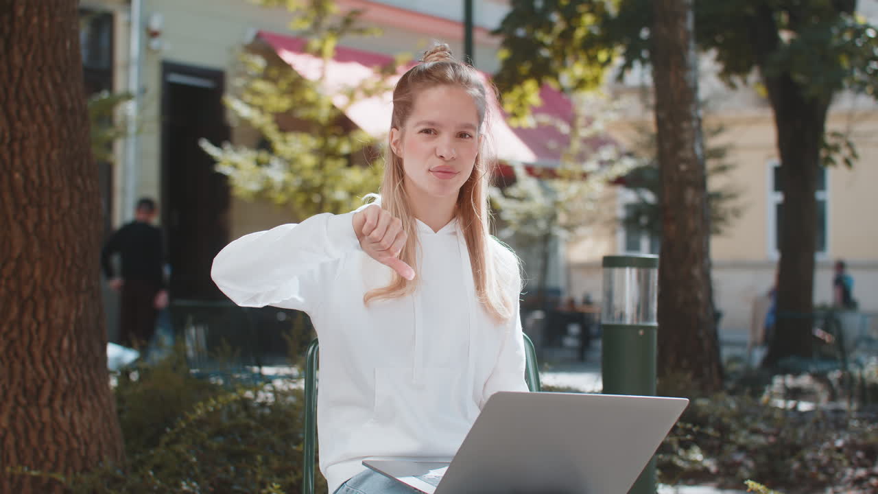 Upset young woman lady using laptop looking at camera and showing thumb down in city street outdoors