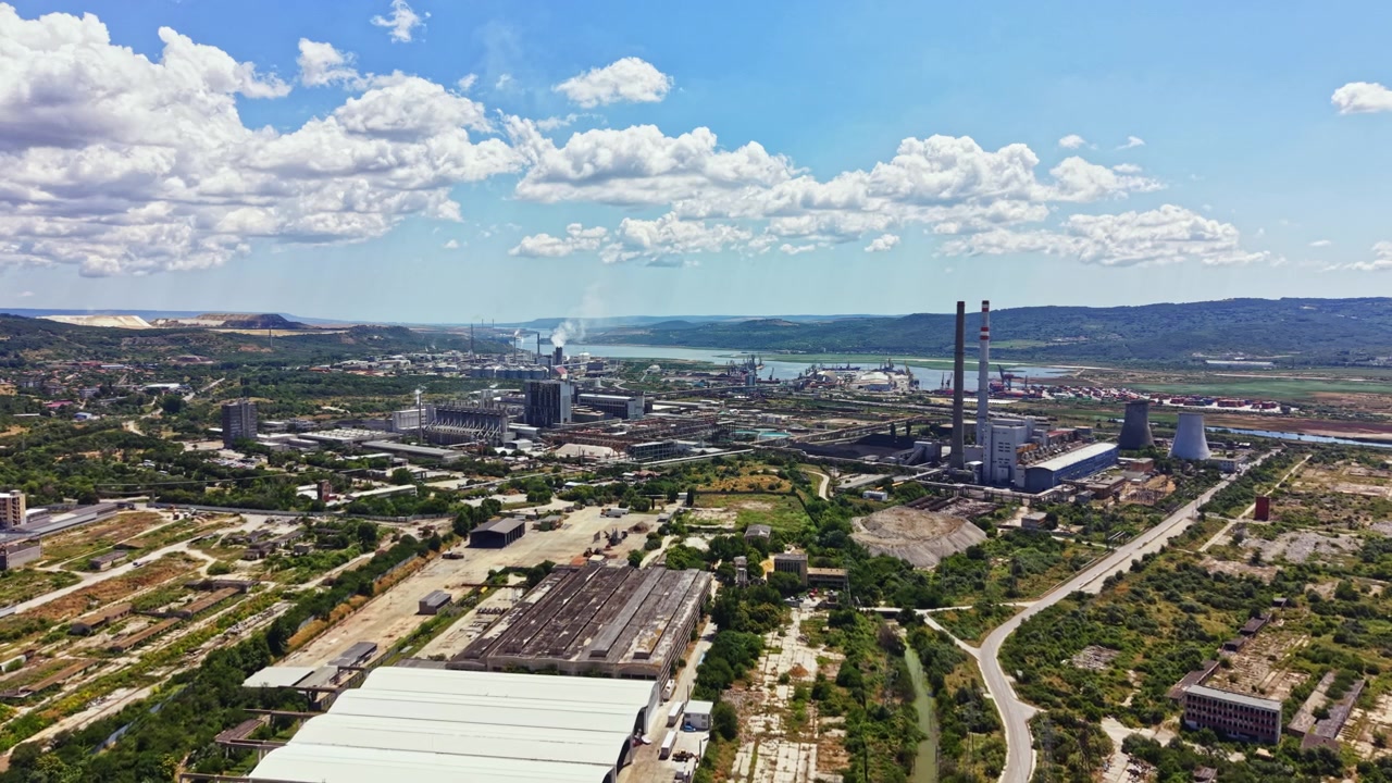 Zona industrial cerca de un río con fábricas, chimeneas y colinas distantes durante el día, con nubes en el cielo.