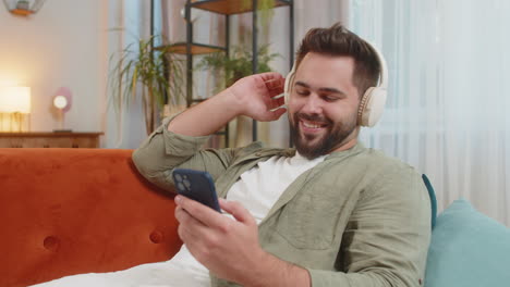 joven feliz y encantado con auriculares inalámbricos usando un teléfono inteligente relajándose sentado en el sofá en casa