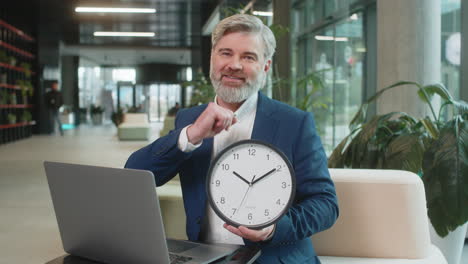 mature businessman with laptop in lobby showing time on wall clock and pointing the finger at camera