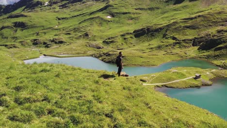 Tiro-De-Paralaje-Aéreo:-Excursionista-De-Pie-En-La-Cima-De-Una-Cresta-De-Montaña-Verde-Con-Vistas-Al-Sendero-Del-Lago-Turquesa-Alpino-De-Bachalpsee-Y-Montañas-Alpinas-Suizas-En-El-Fondo,-Grindelwald,-Suiza-En-Verano