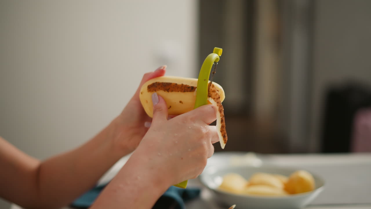 Close up hand view of fair skinned person peeling potato over bowl with skins, kitchen rag nearby, soft blur background of open door and countertop, showing careful home prep and realistic detail