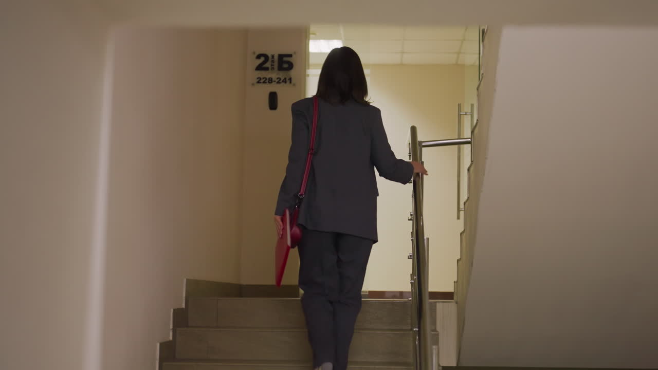 Businesswoman walking upstairs with red leather handbag and notebook, heading to work. Confident and professional in corporate office, symbolizing motivation and focus in career journey