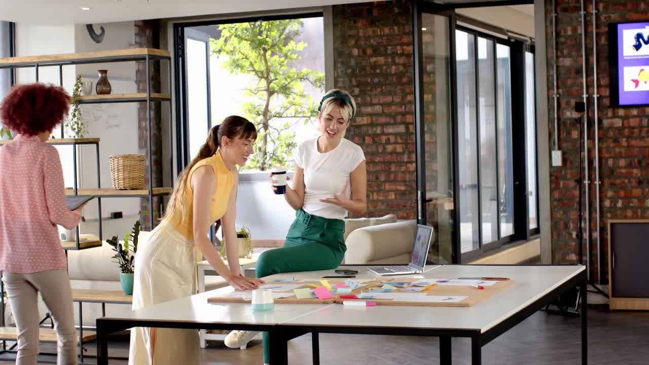 Collaborating in modern office, women discussing ideas with laptop and coffee cup