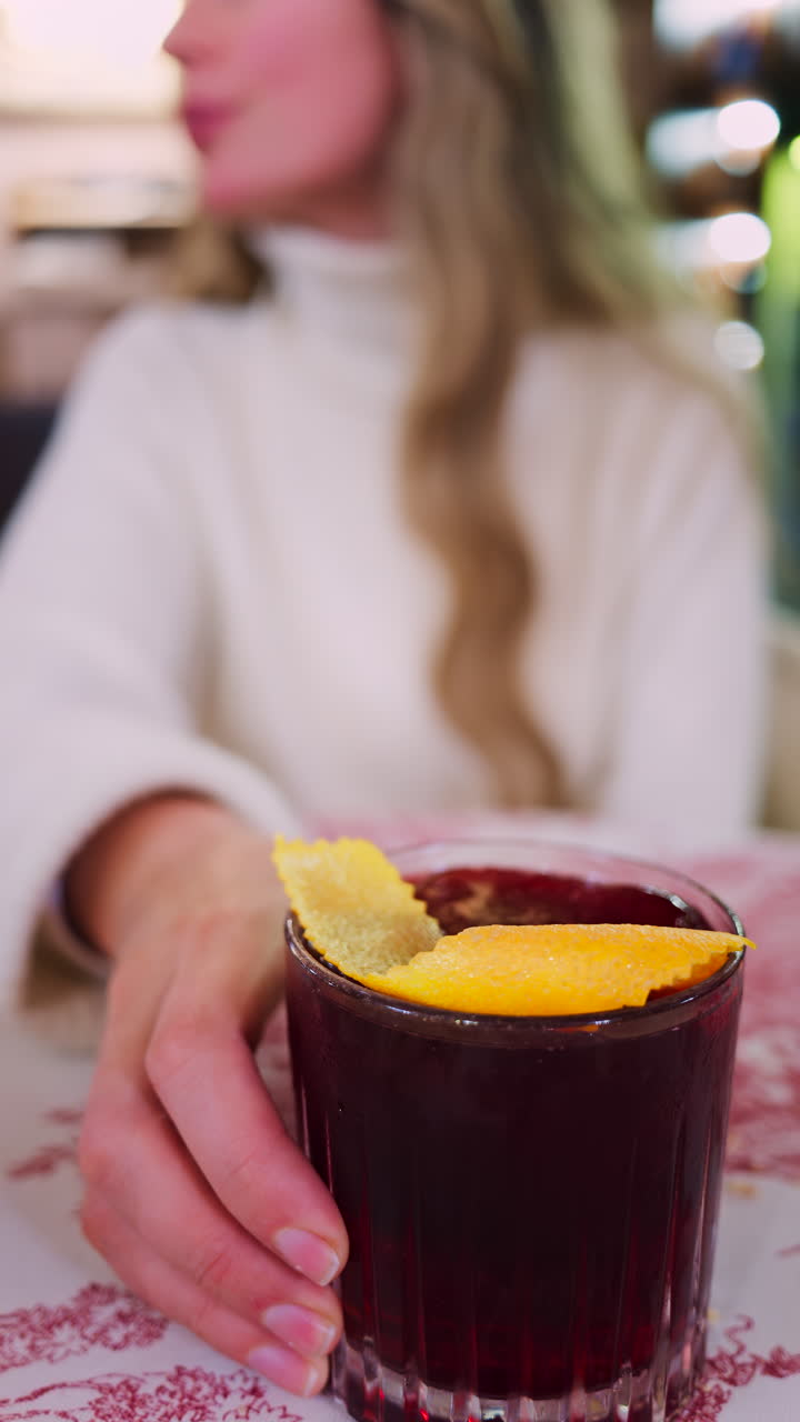 Close up of a woman holding a negroni cocktail on a red and white tablecloth at a restaurant. Vertical